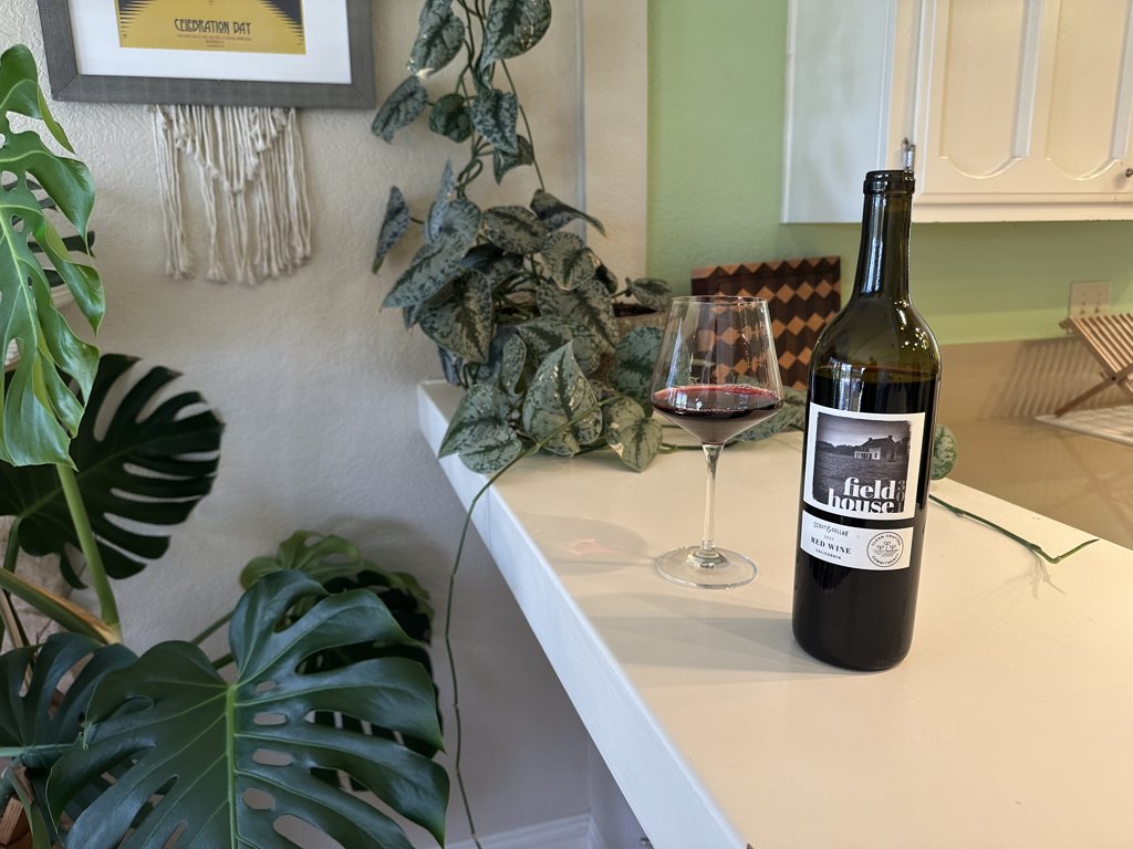 Bottle of 2023 Fieldhouse 301 Red Blend and a glass of red wine on a counter with plants